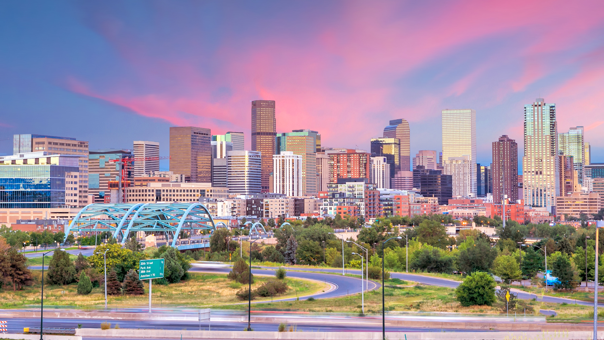 Colorful sunset over the Denver skyline, featuring high-rise buildings, the iconic Millennium Bridge, and surrounding green space in the heart of downtown.