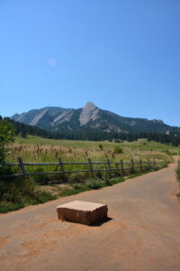 View of the Flatirons from a trail at Chautauqua Park in Boulder, Colorado, with a dirt path, wooden fence, and grassy meadow in the foreground.