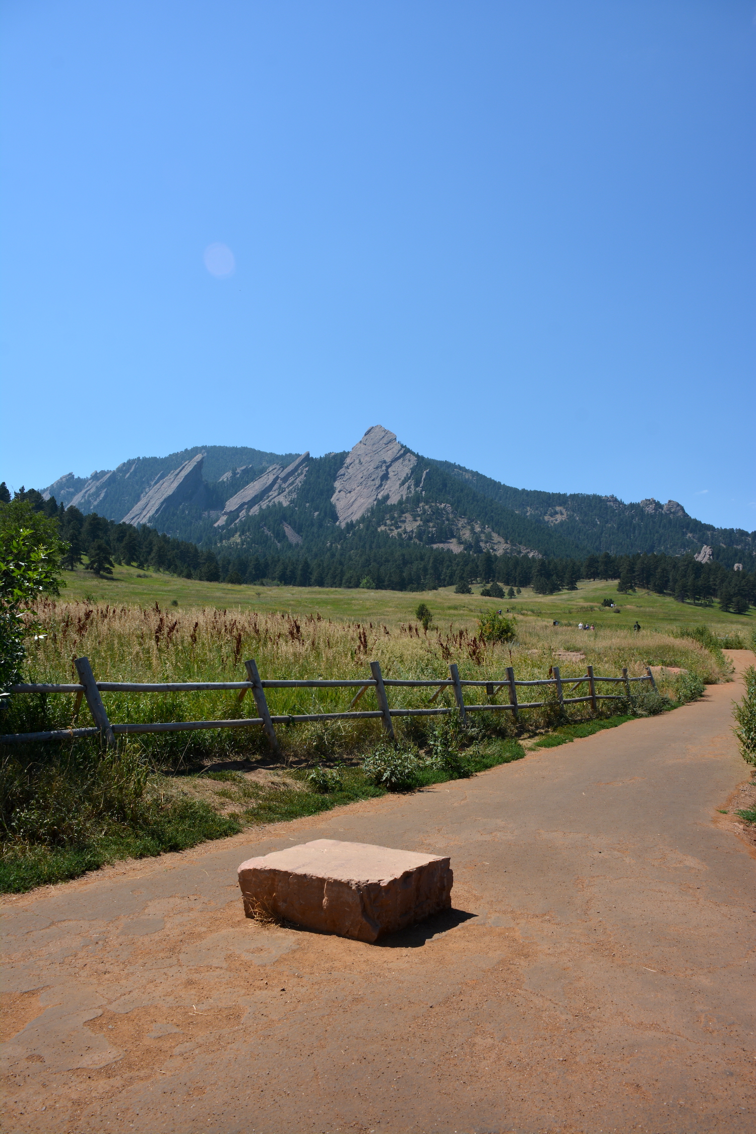 View of the Flatirons from a trail at Chautauqua Park in Boulder, Colorado, with a dirt path, wooden fence, and grassy meadow in the foreground.