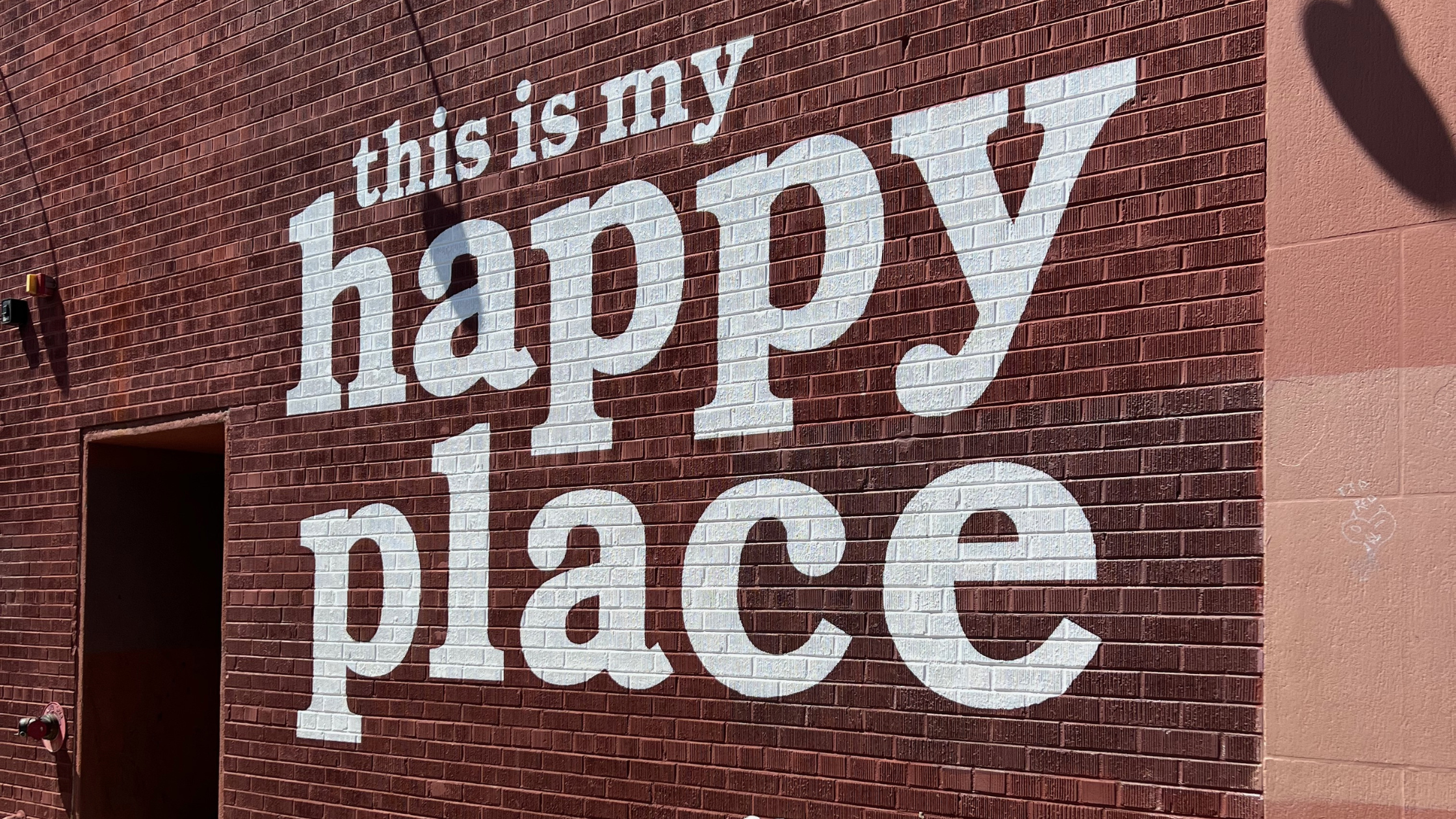 White block letters painted on a red brick wall read “this is my happy place,” part of a mural in Downtown Boulder on the Pearl Street Mall.