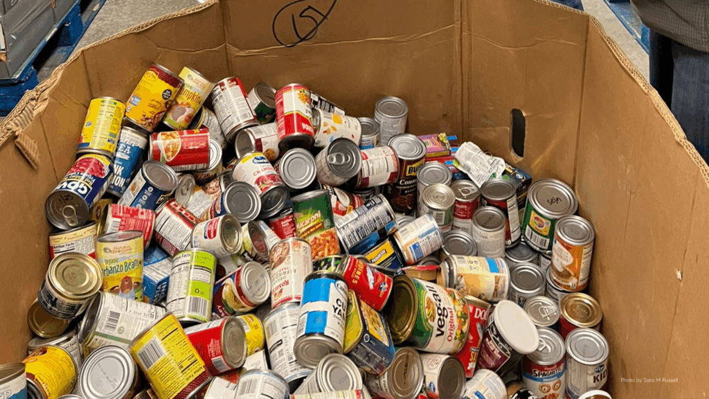 A large cardboard box filled with donated canned goods such as beans, vegetables, soups, and pasta, ready to be sorted and distributed through a community food program.