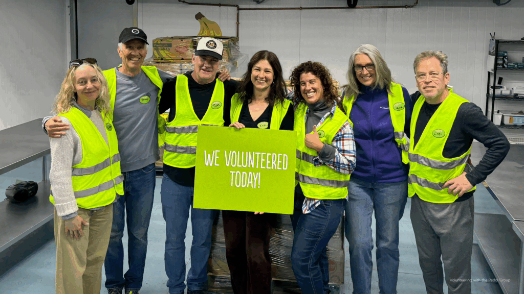 John Farley and several members of The Pedal Group smile together while volunteering at Community Food Share in Boulder County. The team is wearing bright yellow safety vests and holding a green sign that reads “We Volunteered Today!” inside a food distribution area with boxes and supplies in the background.