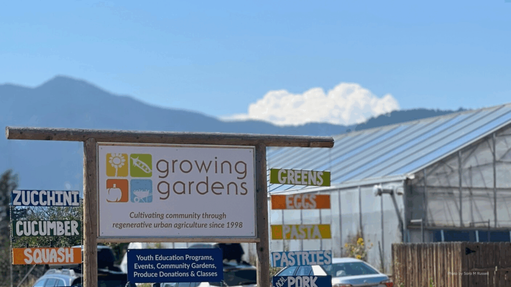 A wooden sign for Growing Gardens stands in front of a greenhouse with the Boulder foothills in the background. The sign reads “Growing Gardens – Cultivating community through regenerative urban agriculture since 1998,” surrounded by colorful smaller signs for local produce like zucchini, cucumber, squash, greens, and eggs.