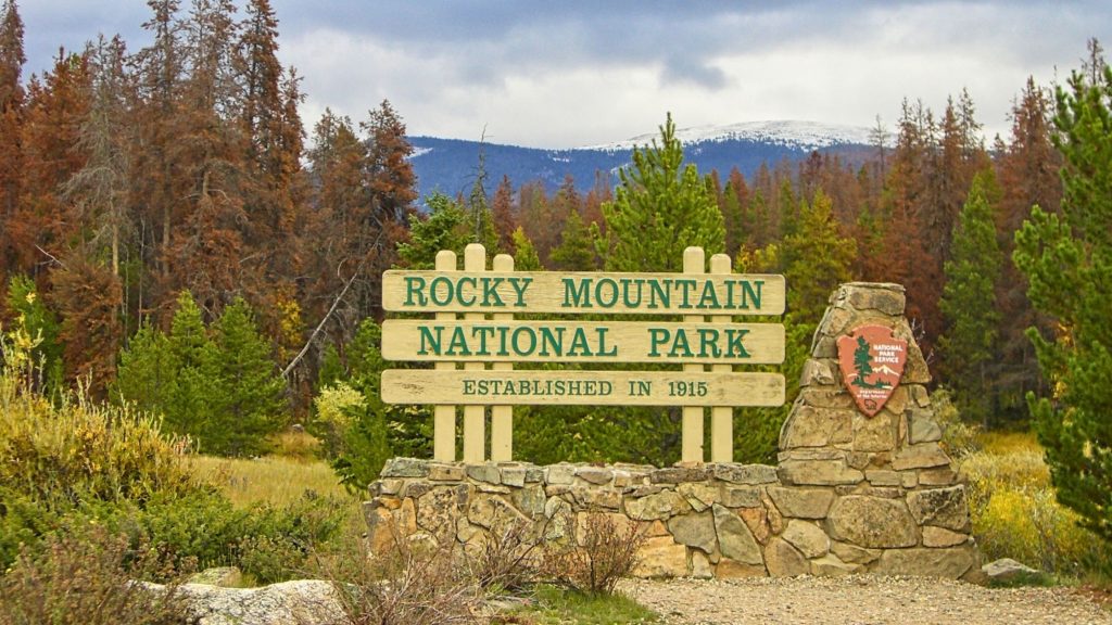 Rocky Mountain National Park entrance surrounded by evergreen trees and early fall foliage with snow visible on the mountains.