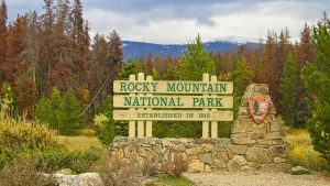 Rocky Mountain National Park entrance surrounded by evergreen trees and early fall foliage with snow visible on the mountains.